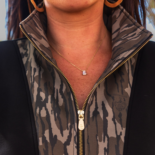 Female wearing a camouflage-patterned outfit with a gold zipper, showing a necklace with a small pendant. Garment is accented in Mossy Oak Bottomland for the camo accent.