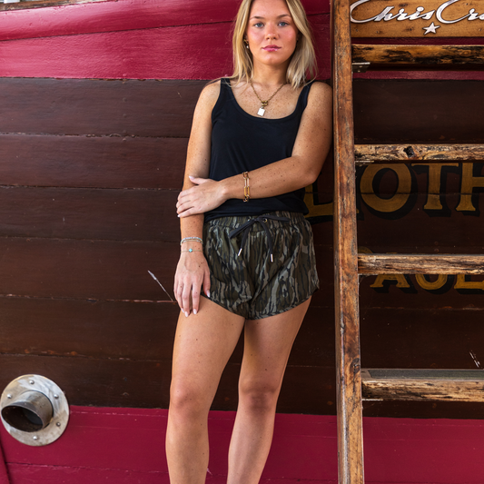 Woman in black tank top and GreeneCo Mossy Oak Bottomland camouflage shorts.