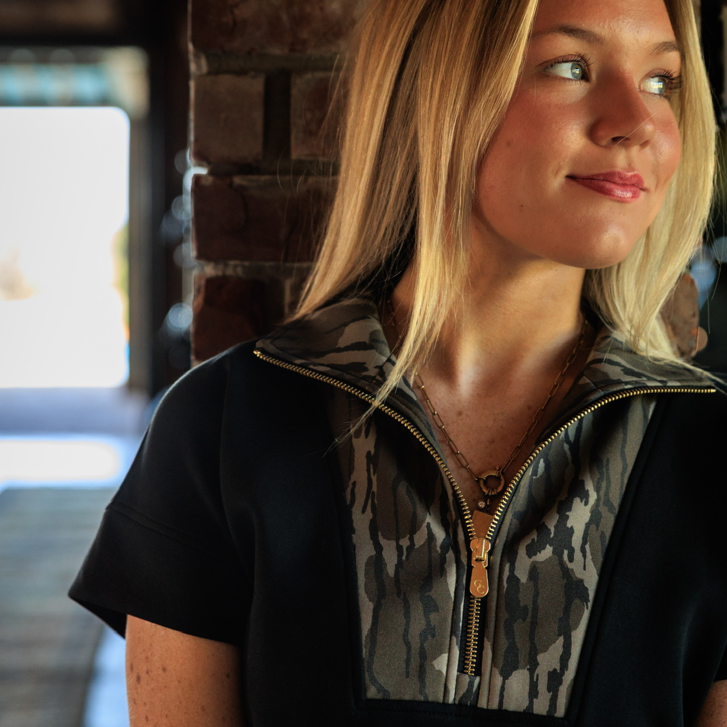 Woman with blonde hair wearing a black and Mossy Oak camouflage garment with a brick wall background. Emphasis on the shiny gold zipper. 