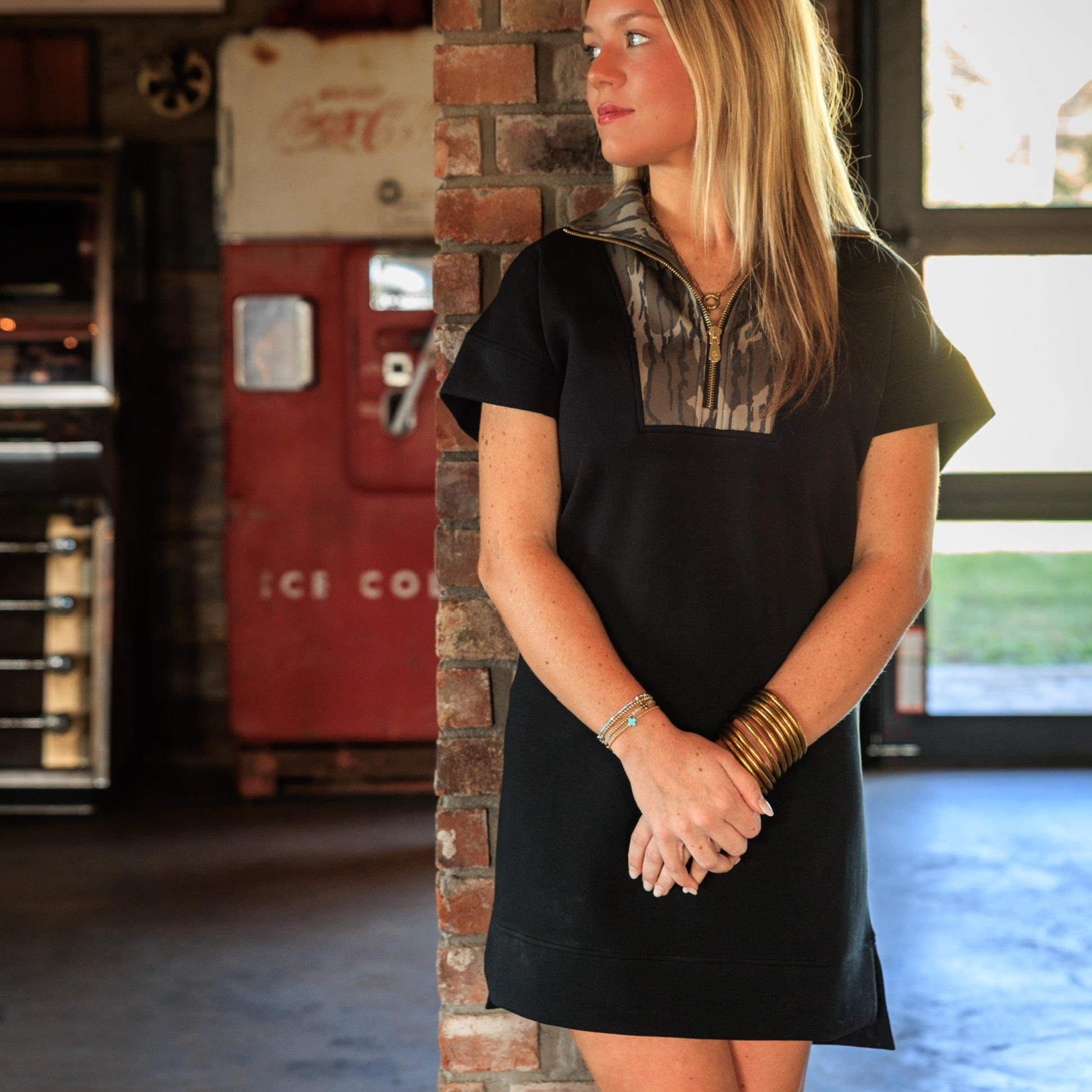 Woman in a black dress with Mossy Oak camouflage accents standing against a brick wall.