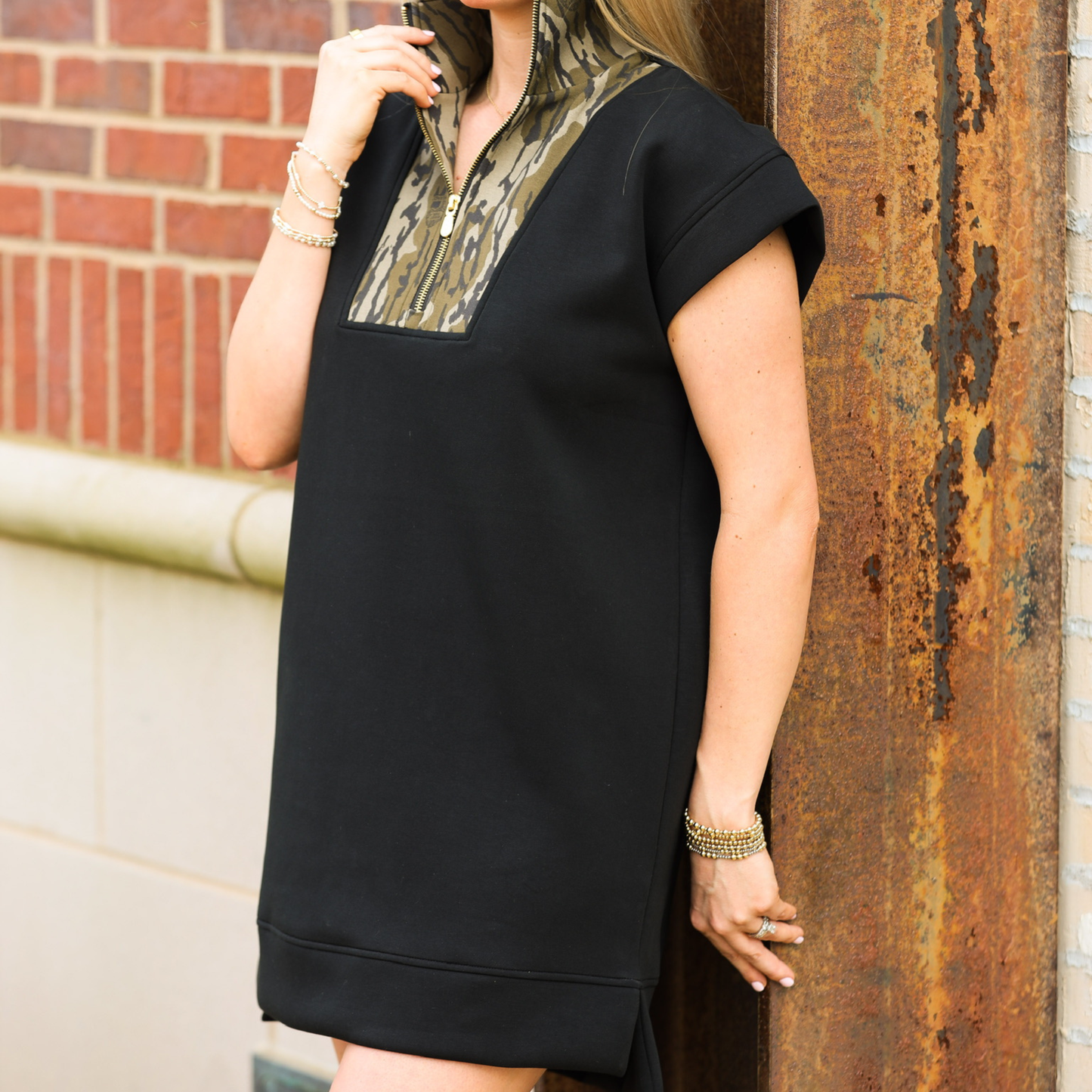 Person wearing a black dress with a camo patterned neckline against a brick wall.