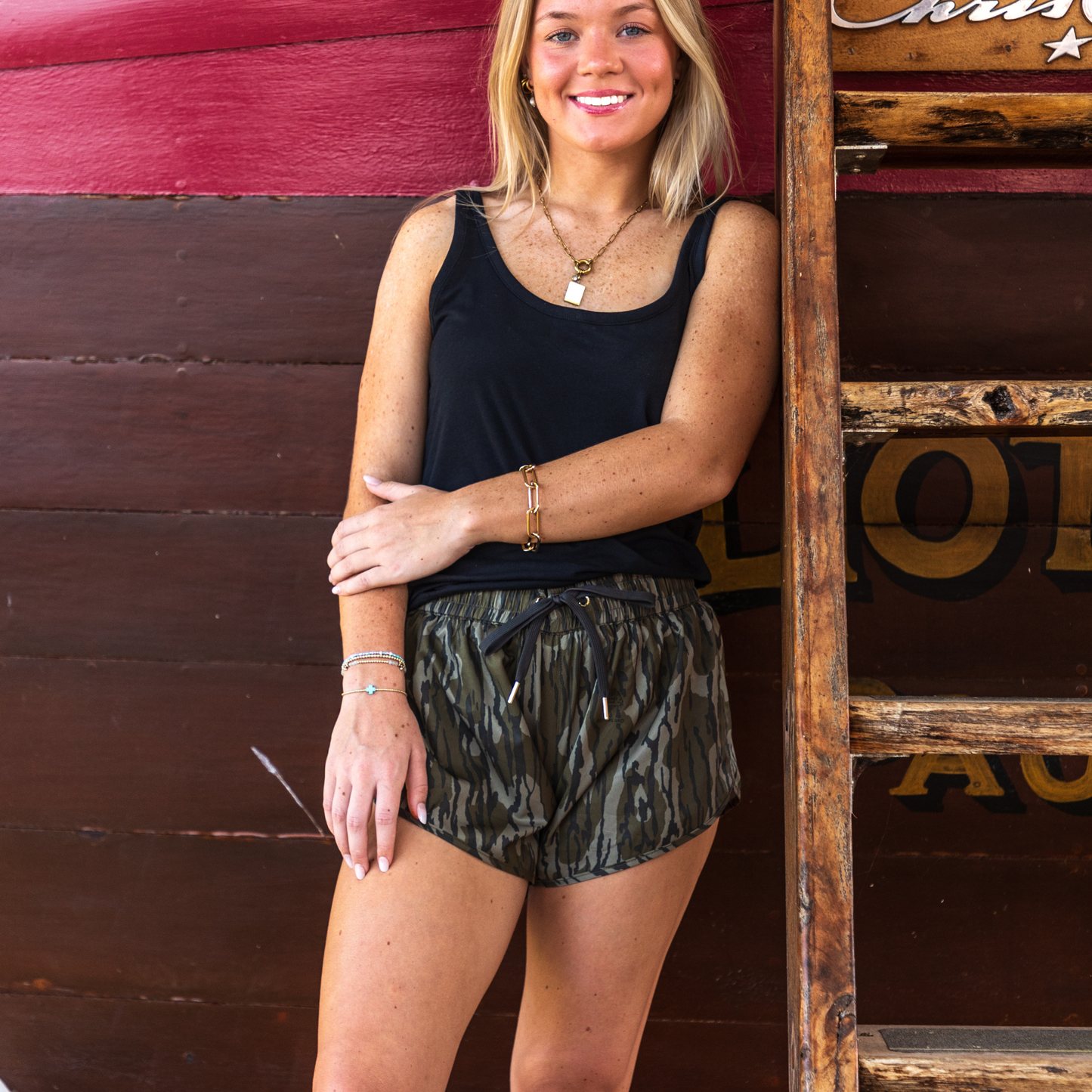 Woman wearing a black tank top and camouflage GreeneCo River shorts standing against a wooden background.