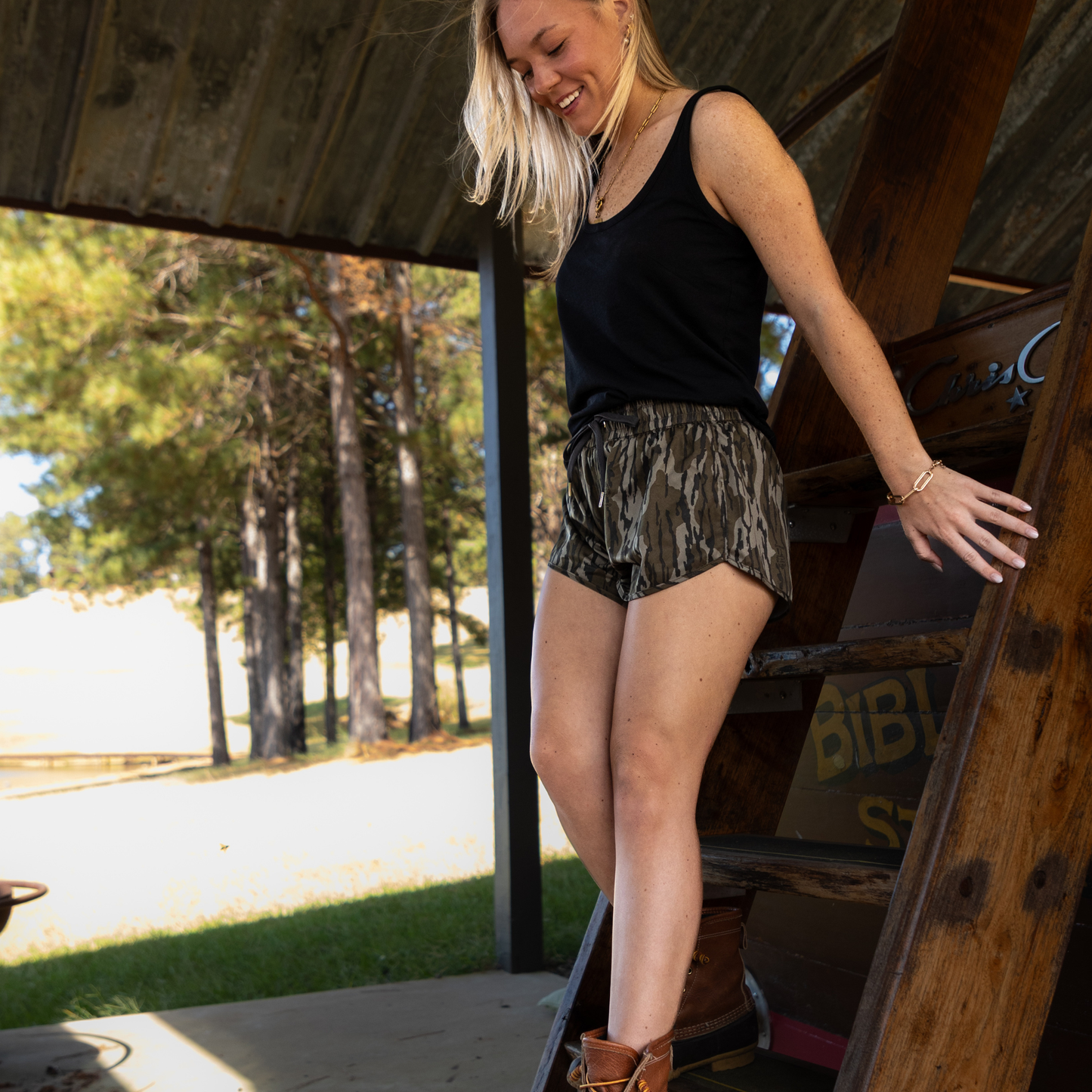 Woman in black tank top and GreeneCo Mossy Oak Bottomland camo athletic shorts standing next to a wooden structure with trees in the background.