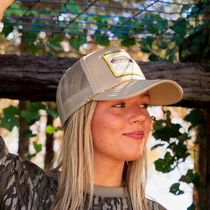 Model styling a beige trucker hat with GreeneCo logo and removable gold chain accent