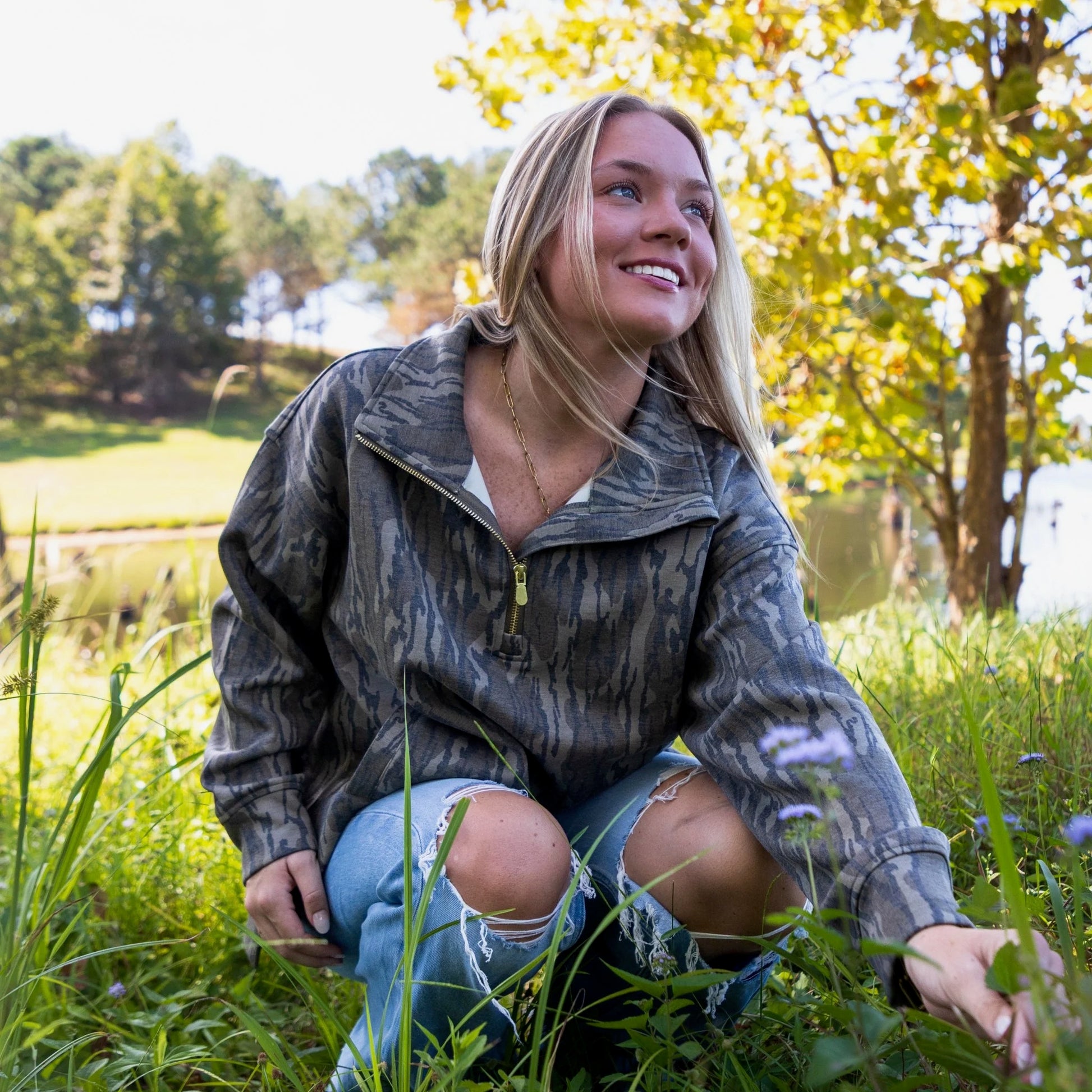 Woman in Mossy Oak camouflage pullover sitting in a grassy field with trees and water to showcase versatility of the garment going from outdoor use to every day athleisure . 