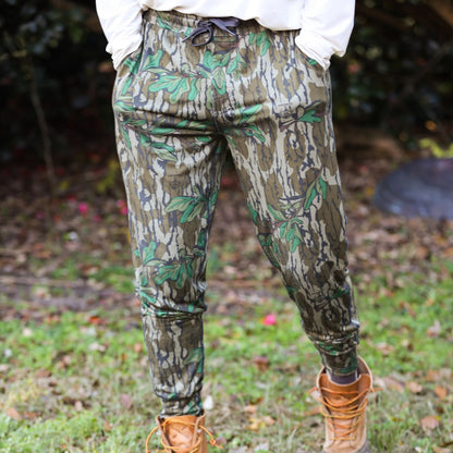 Camouflage jogger style pants worn outdoors with a blurred natural background to showcase athletic fit, tapered leg and elastic drawstring for the waistband. 