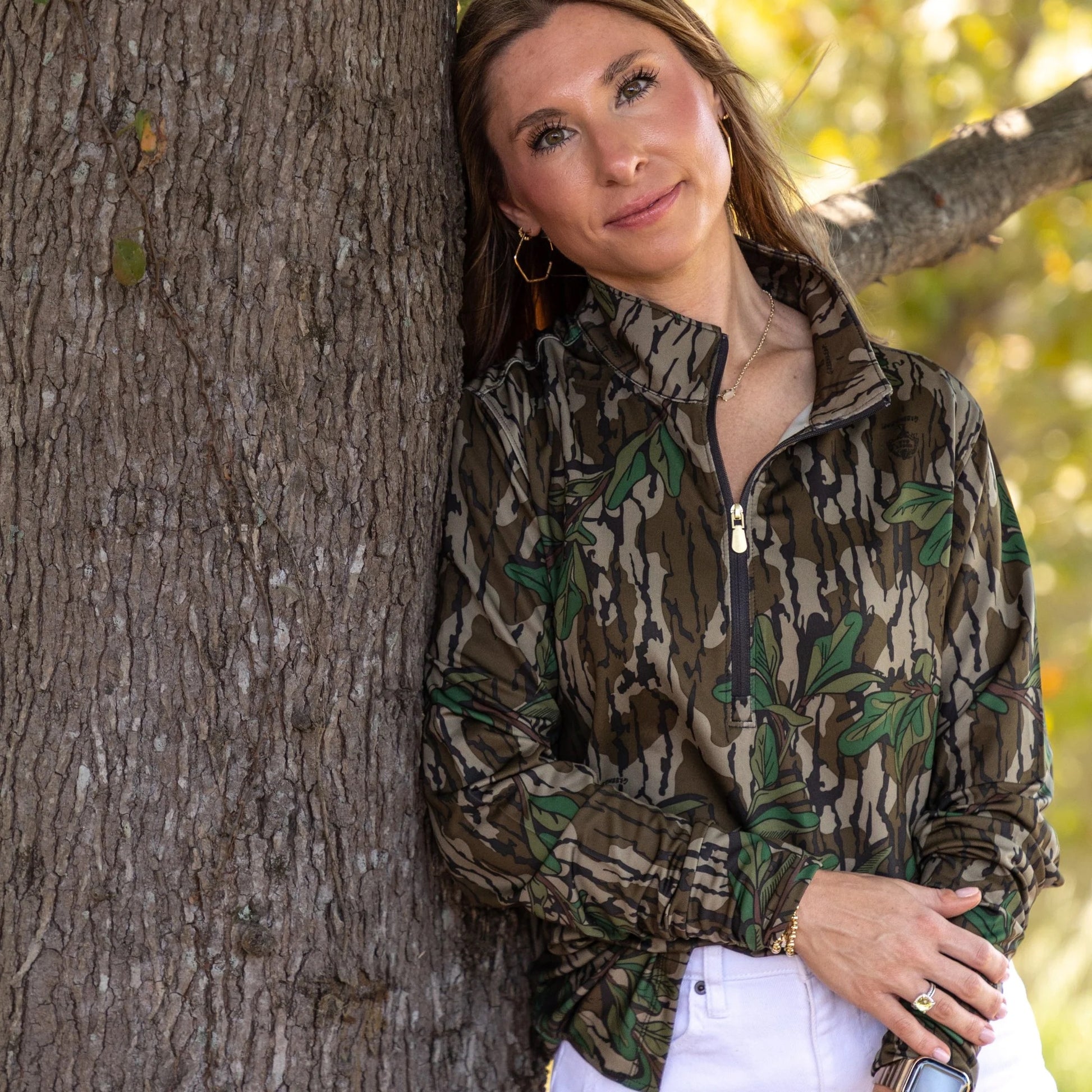 Women wearing athleisure camouflage paired with white shorts to offer versatility in styling. Gold zipper accent on the half-zip style showcasing the Mossy Oak greenleaf pattern.