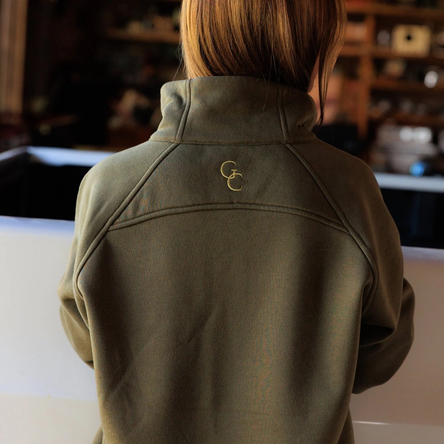 Women’s olive pullover back view to highlight the structure and fit of the pullover garment. Close up detail of drop shoulder style detail and a stacked embroidered logo in gold threading. 