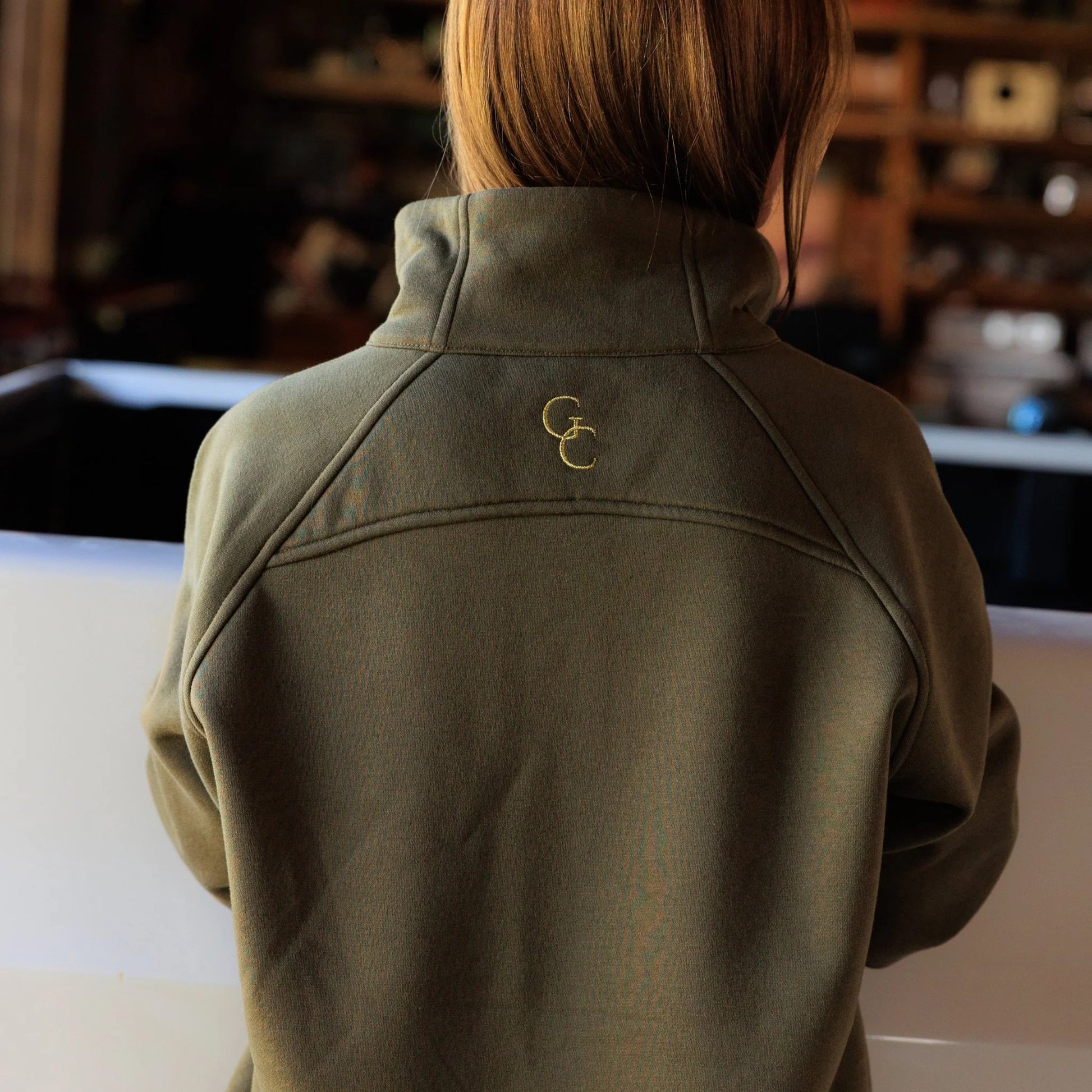 Women’s olive pullover back view to highlight the structure and fit of the pullover garment. Close up detail of drop shoulder style detail and a stacked embroidered logo in gold threading. 
