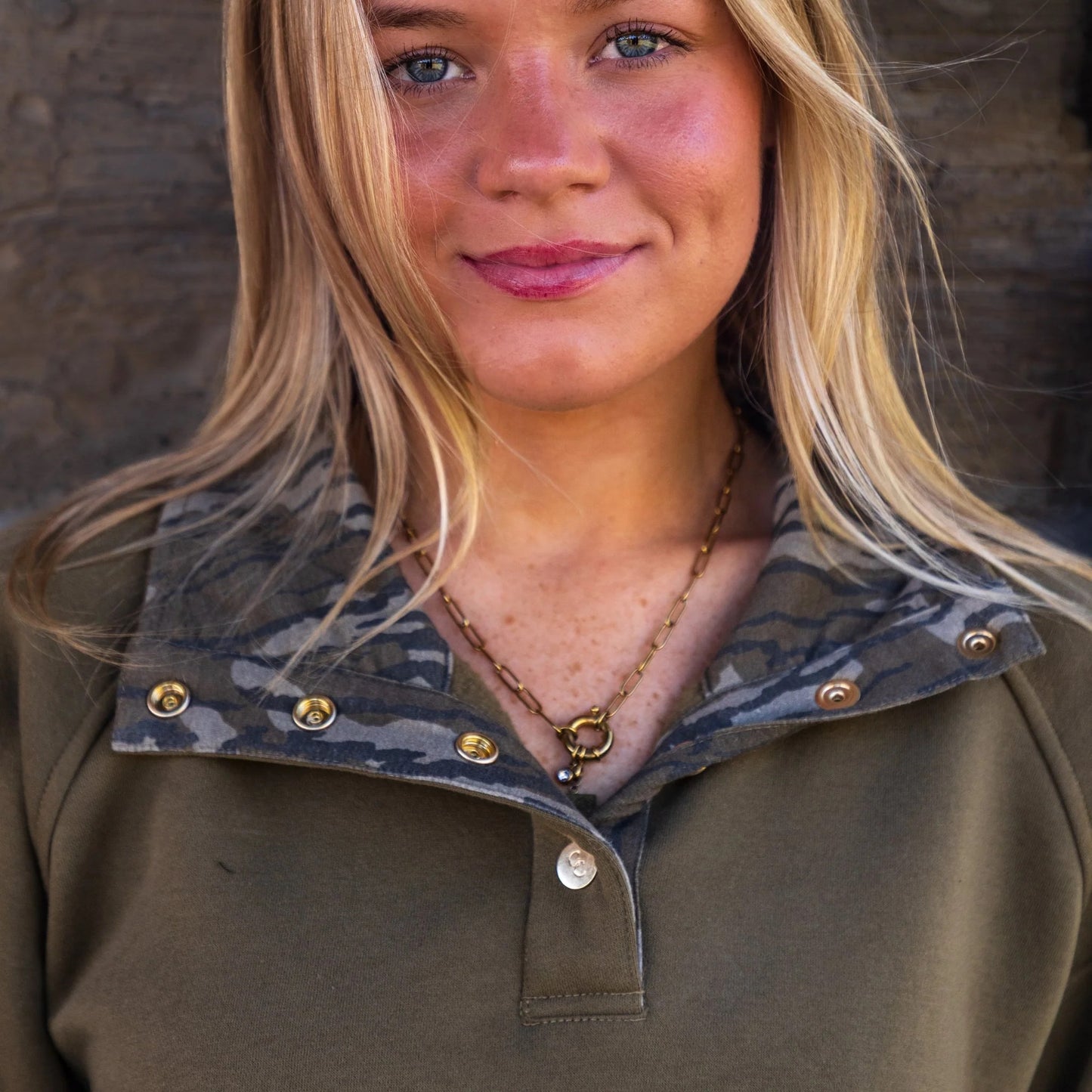 Model wearing olive quarter-snap pullover showcasing Mossy Oak Bottomland camouflage around the interior collar highlighting the elevated gold snap button detail. 