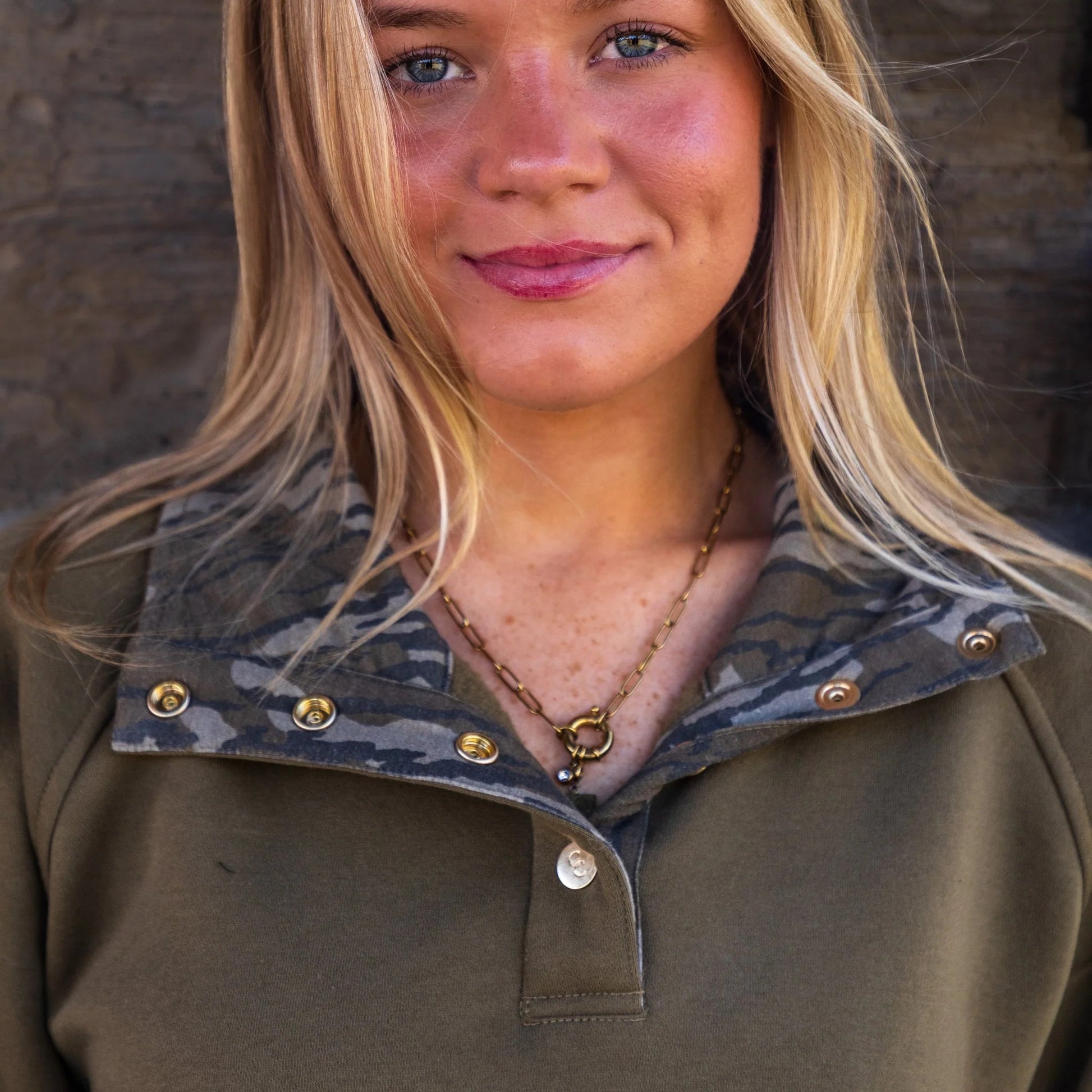 Model wearing olive quarter-snap pullover showcasing Mossy Oak Bottomland camouflage around the interior collar highlighting the elevated gold snap button detail. 